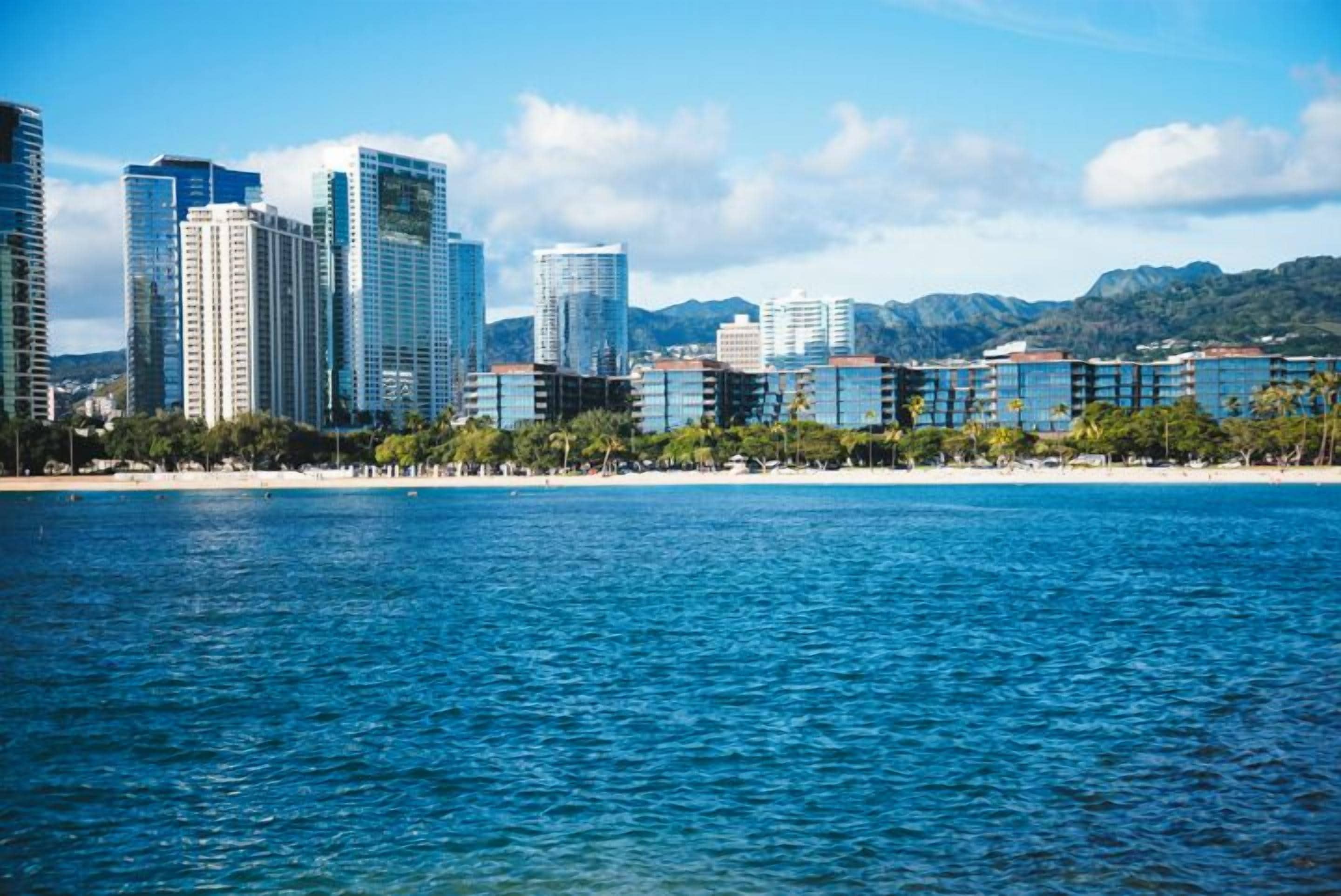 A coastal cityscape with modern high-rise buildings, a beach, and mountains in the background.