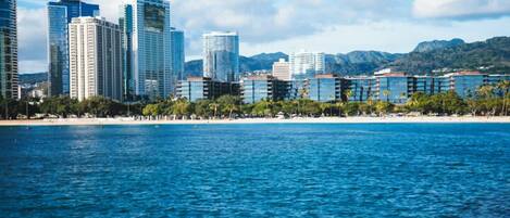 A coastal cityscape with modern high-rise buildings, a beach, and mountains in the background.