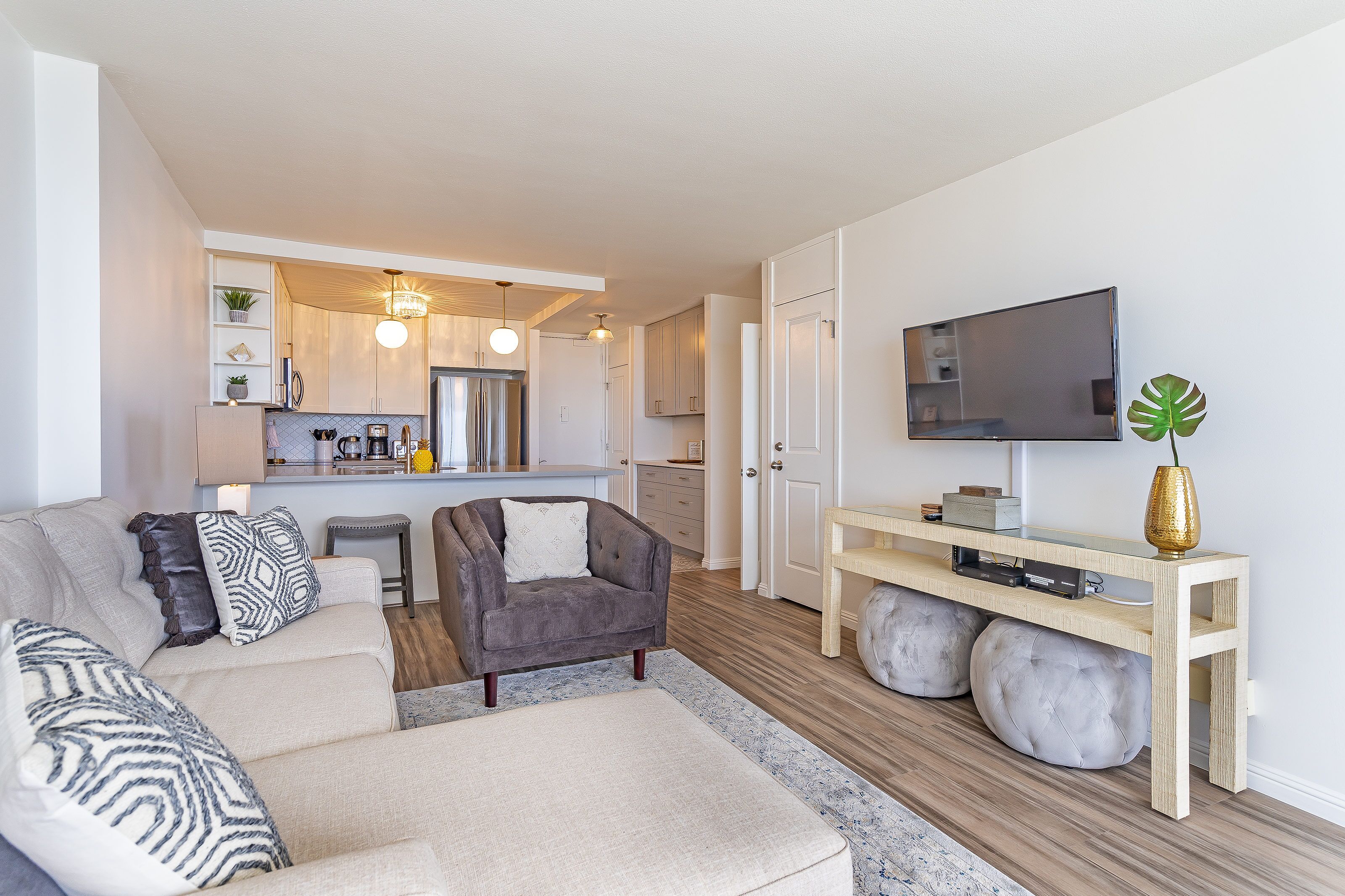 A modern living room with a sofa, armchair, and ottoman, a TV on a stand, and a kitchen in the background.