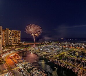 Un port de plaisance avec des bateaux amarrés, un grand bâtiment doté de nombreuses fenêtres et des feux d’artifice dans le ciel.