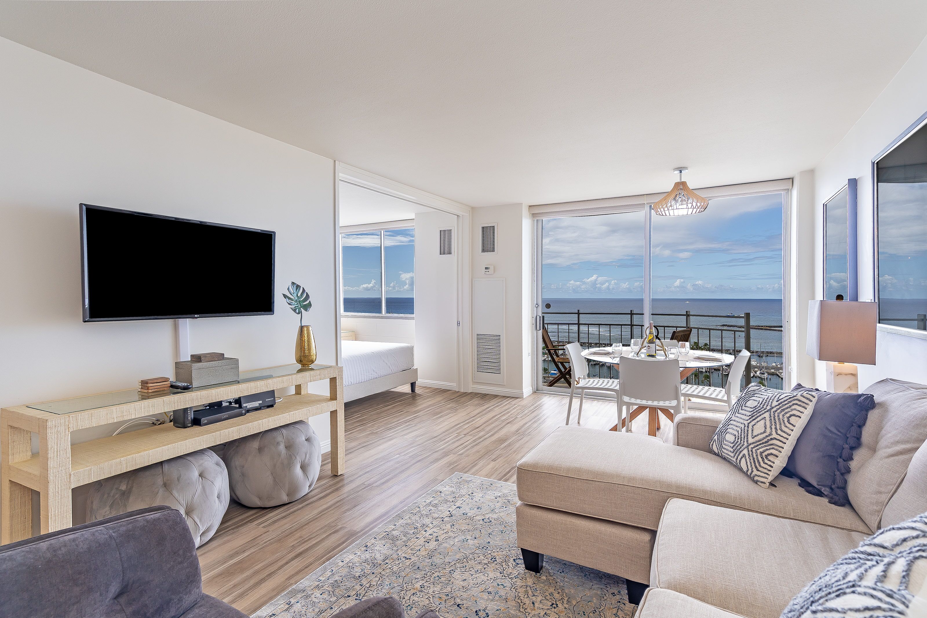 A modern living room with a flat-screen TV, a sofa, a coffee table, and a balcony with a view of the sea.