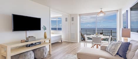 A modern living room with a flat-screen TV, a sofa, a coffee table, and a balcony with a view of the sea.