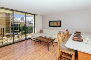A modern living room with a sofa, dining table, chairs, and a balcony with a view of the city. - Ocean-city Views Family Getaway at Waikiki Banyan Half a Block to the Beach (Honolulu)