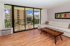 A modern living room with a white sofa, a wooden coffee table, and a large sliding glass door leading to an outdoor patio with a table and chairs. - Ocean-city Views Family Getaway at Waikiki Banyan Half a Block to the Beach (Honolulu)