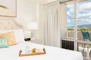 Ein Hotelzimmer mit Bett, Nachttisch mit Lampe, Tablett mit Tasse und Obst sowie Blick auf einen Balkon mit Stuhl.