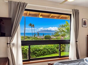 A room with a large window offering a view of a tropical landscape, including palm trees and a mountain. - Maui Kaʻanapali Condo - Ocean View, Walk to Beach (Lahaina)