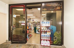 A store entrance with a glass door, shelves stocked with bottled water, and a sign indicating the store is open 8am. - Ocean & City Views at Island Colony W/full Kitchen & Close to the Beach (Honolulu)