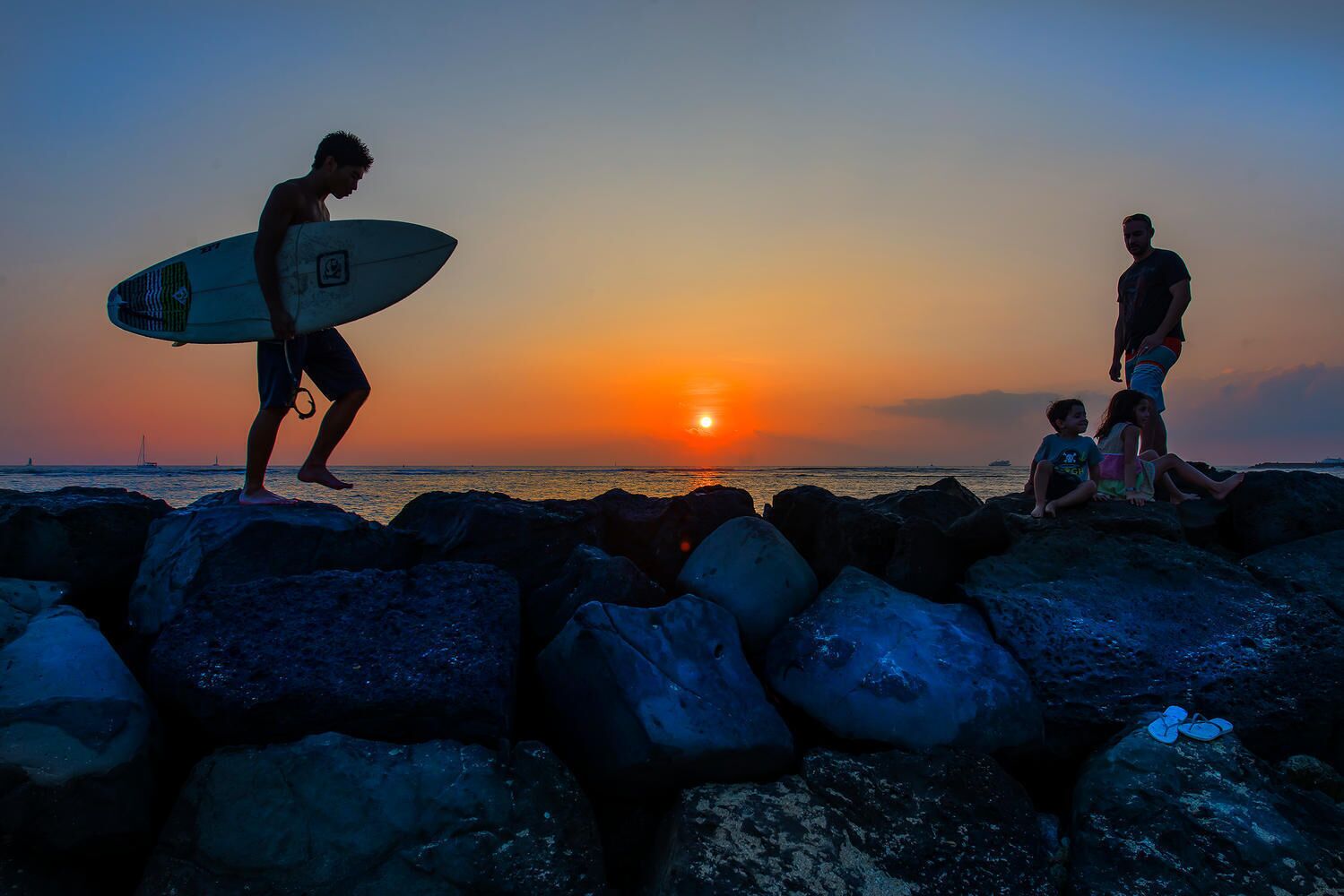 Escena al atardecer con una persona llevando una tabla de surf y otras personas sentadas sobre rocas junto al agua.