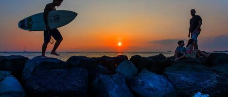 Escena al atardecer con una persona llevando una tabla de surf y otras personas sentadas sobre rocas junto al agua.