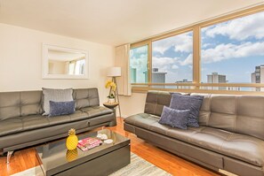 A modern living room with two dark leather sofas, a glass coffee table, and a view of the city skyline through large windows. - Ocean & City Views at Island Colony W/full Kitchen & Close to the Beach (Honolulu)