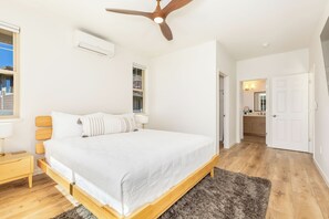 Ein Schlafzimmer mit Bett, Holz-Kopfteil, Nachttischen, Deckenventilator, Klimaanlage, Fenster und einer Tür zu einem Badezimmer.