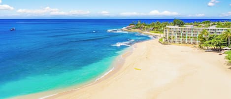 A beach with clear turquoise waters, a sandy shore, and a hotel with multiple floors and balconies overlooking the ocean.