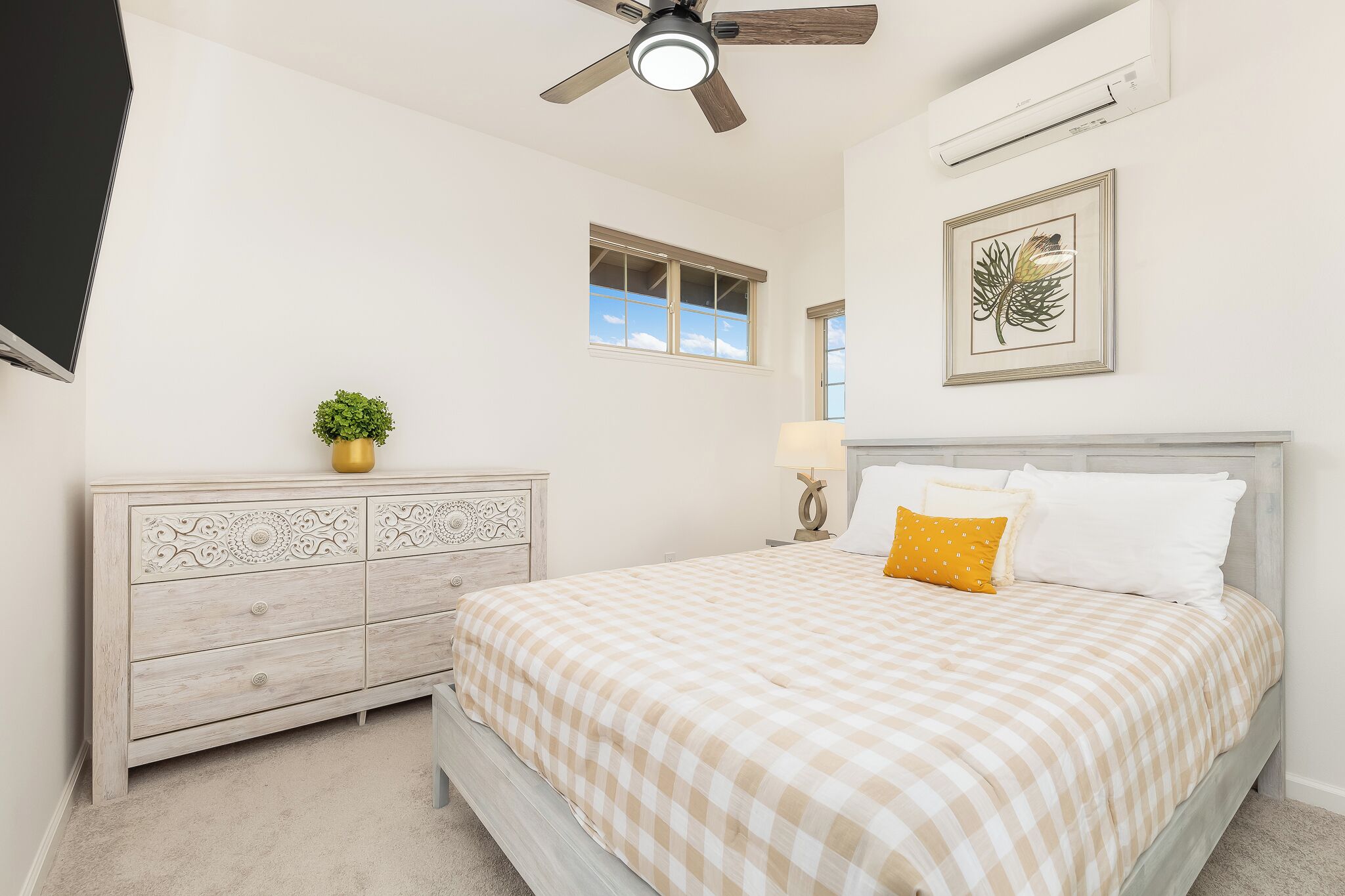 Une chambre à coucher comprenant un lit, une commode, un ventilateur de plafond, une fenêtre et des œuvres d’art.