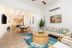 A modern living room with a round coffee table, a sofa with beach-themed cushions, a dining area with wooden chairs, and a kitchen with a flat-screen TV. - Ocean View Home for Family Fun Getaway Near Beach W/hot Tub and Bbq Grill (Waianae)