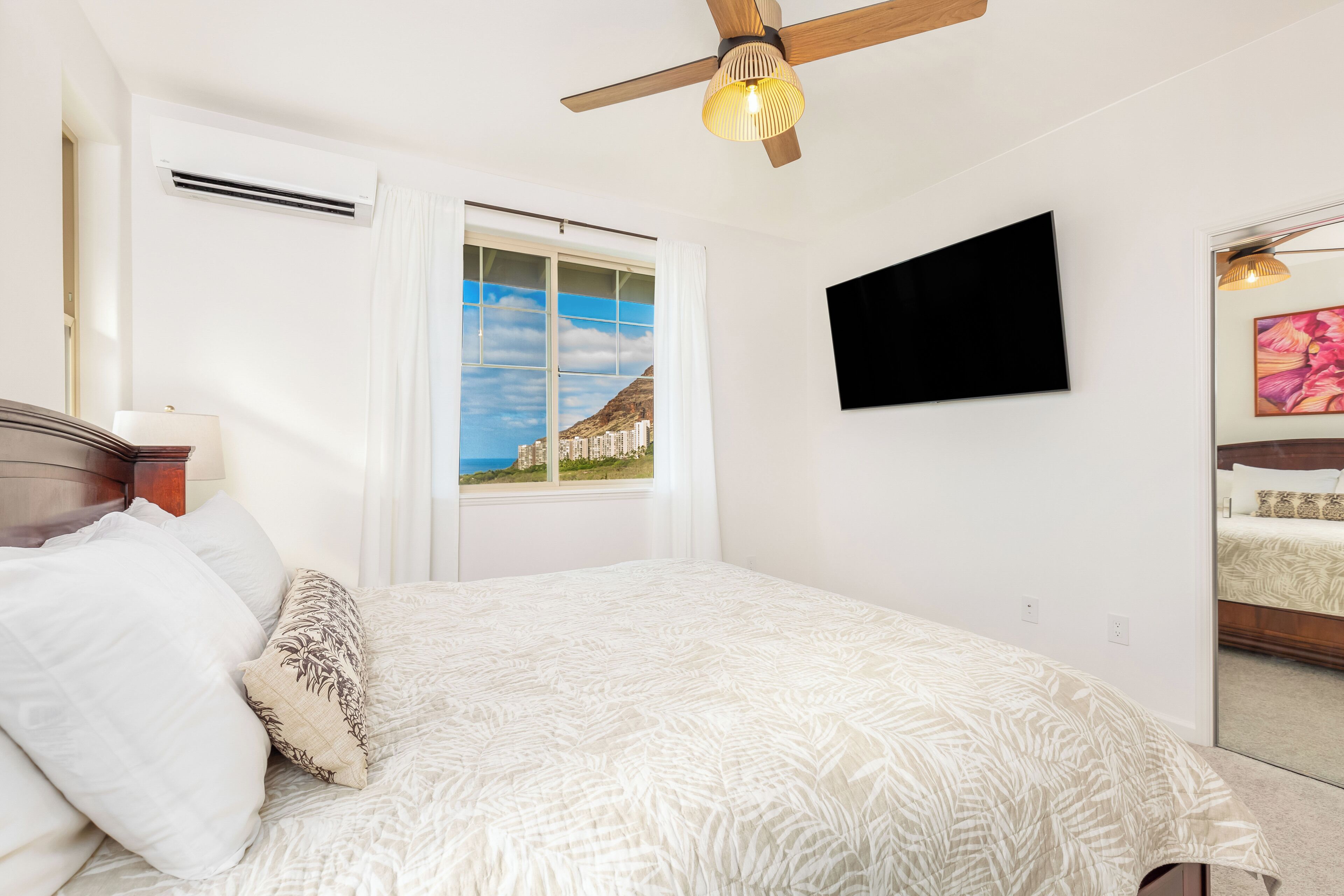 A bedroom with a bed, a ceiling fan, a window with a view, a flat-screen TV, and a painting on the wall.