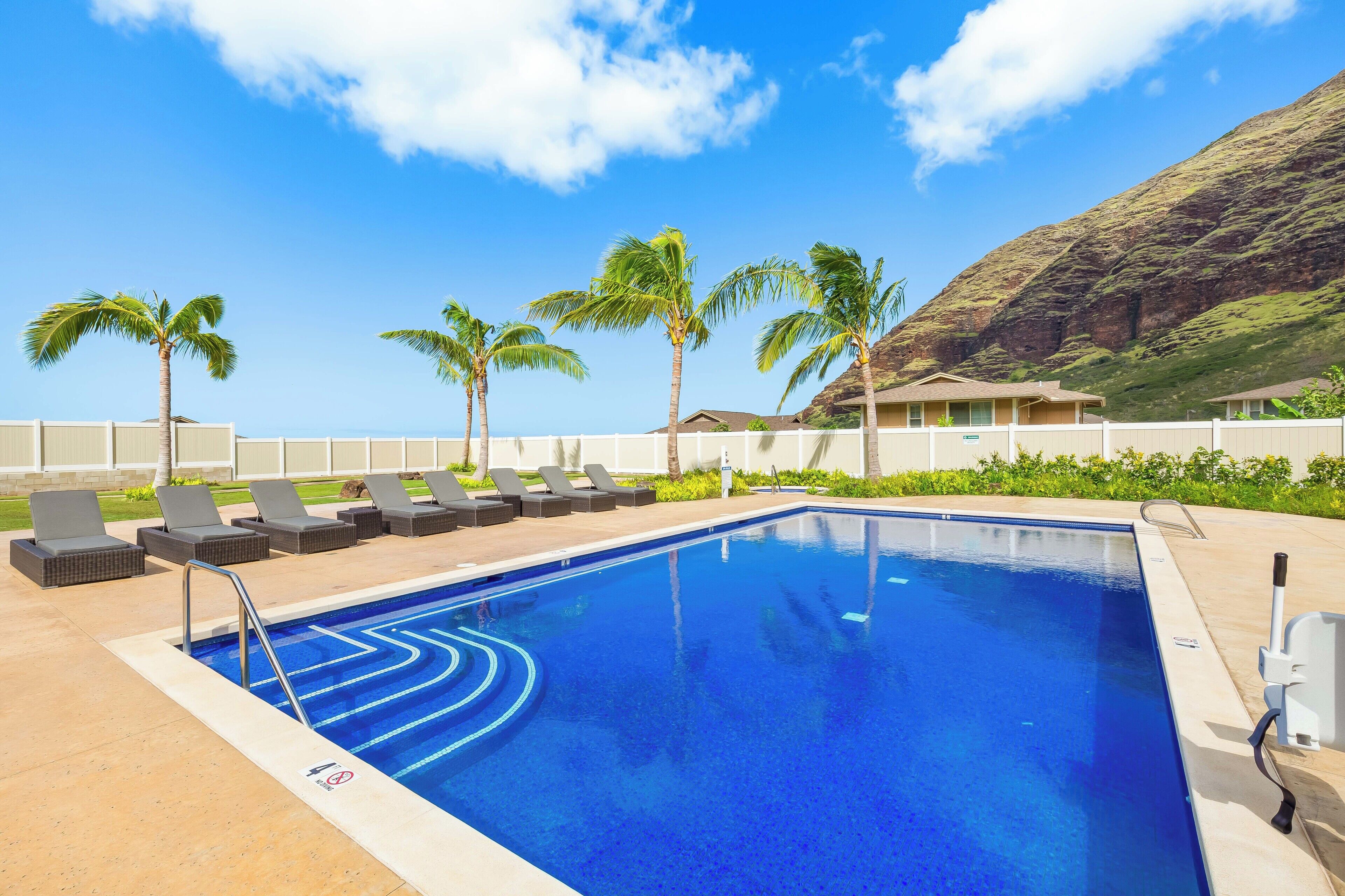 A swimming pool with lounge chairs and palm trees, surrounded by a white fence and a mountain in the background.