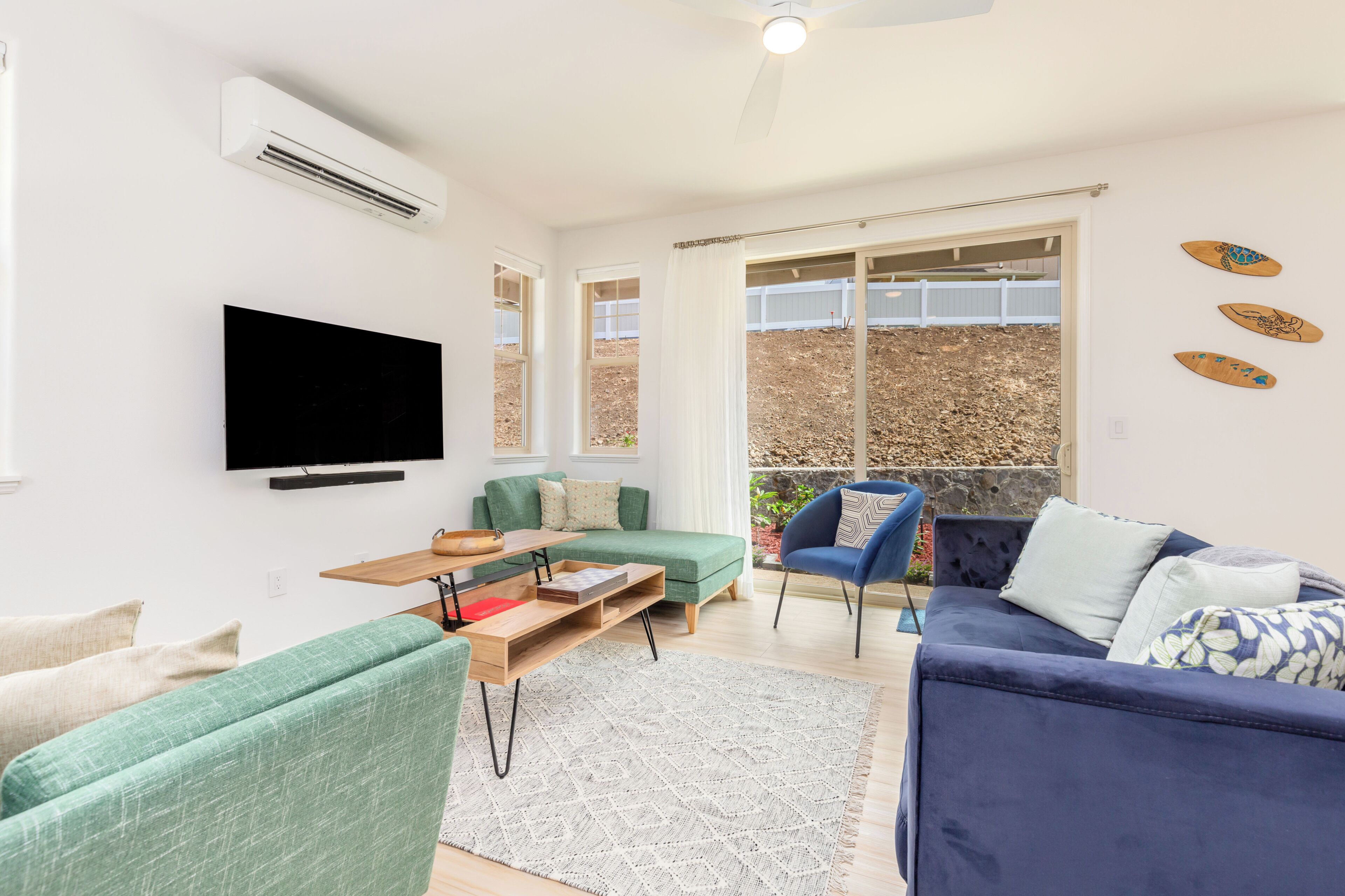 A modern living room with a flat-screen TV, green and blue sofas, a wooden coffee table, and a large window with a view of a garden.