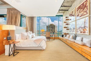 Ein modernes Hotelzimmer mit einem großen Bett, einem Schreibtisch und Blick auf eine Stadtkulisse durch raumhohe Fenster.