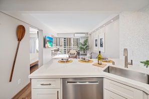 A modern kitchen with a stainless steel dishwasher, a white countertop, and a view of the balcony. - Ocean-lagoon Condo W/full Kitchen at Ilikai Hotel and Steps to the Beach (Honolulu)