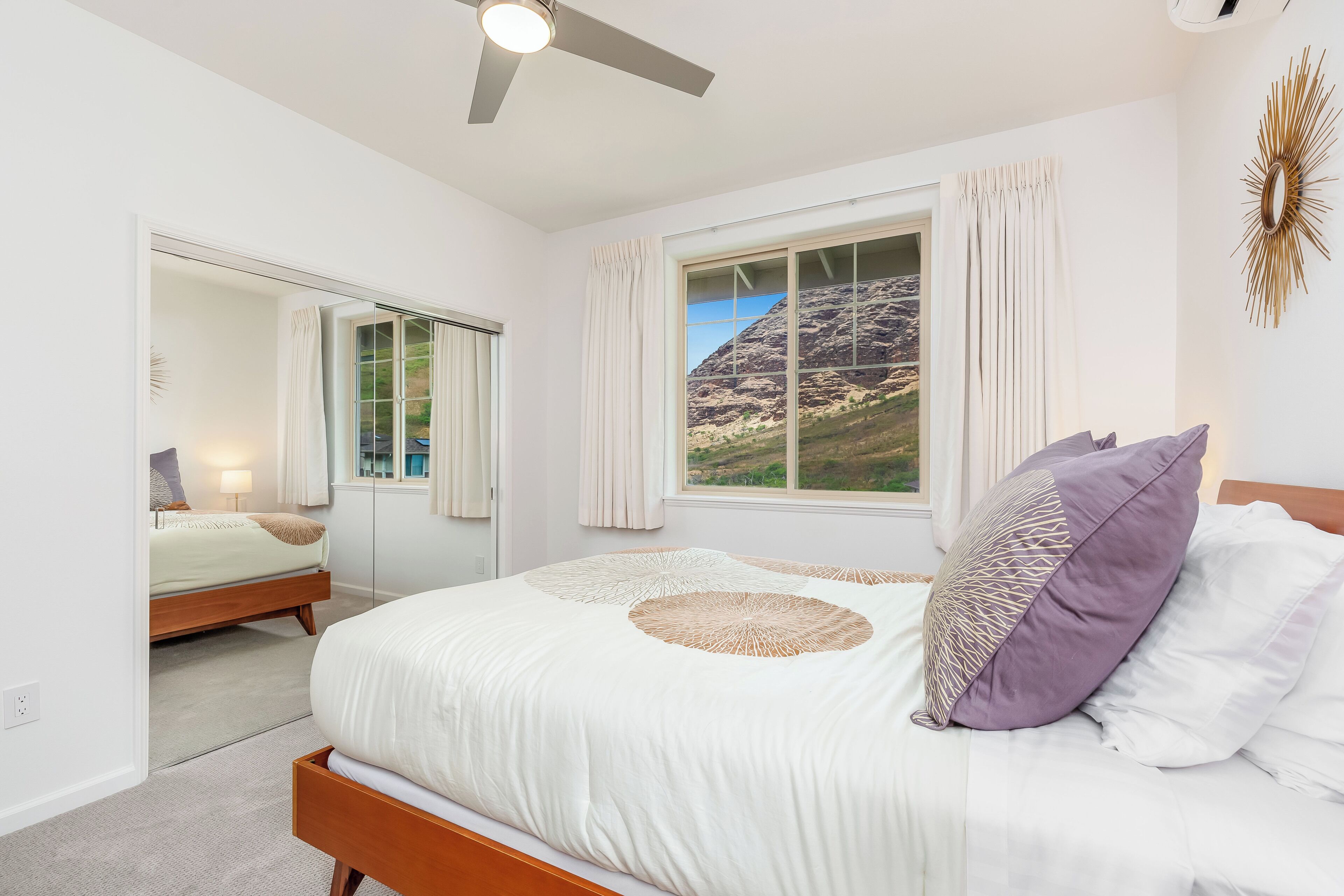 Een slaapkamer met een bed, een raam met gordijnen, een plafondventilator en uitzicht op een berg.