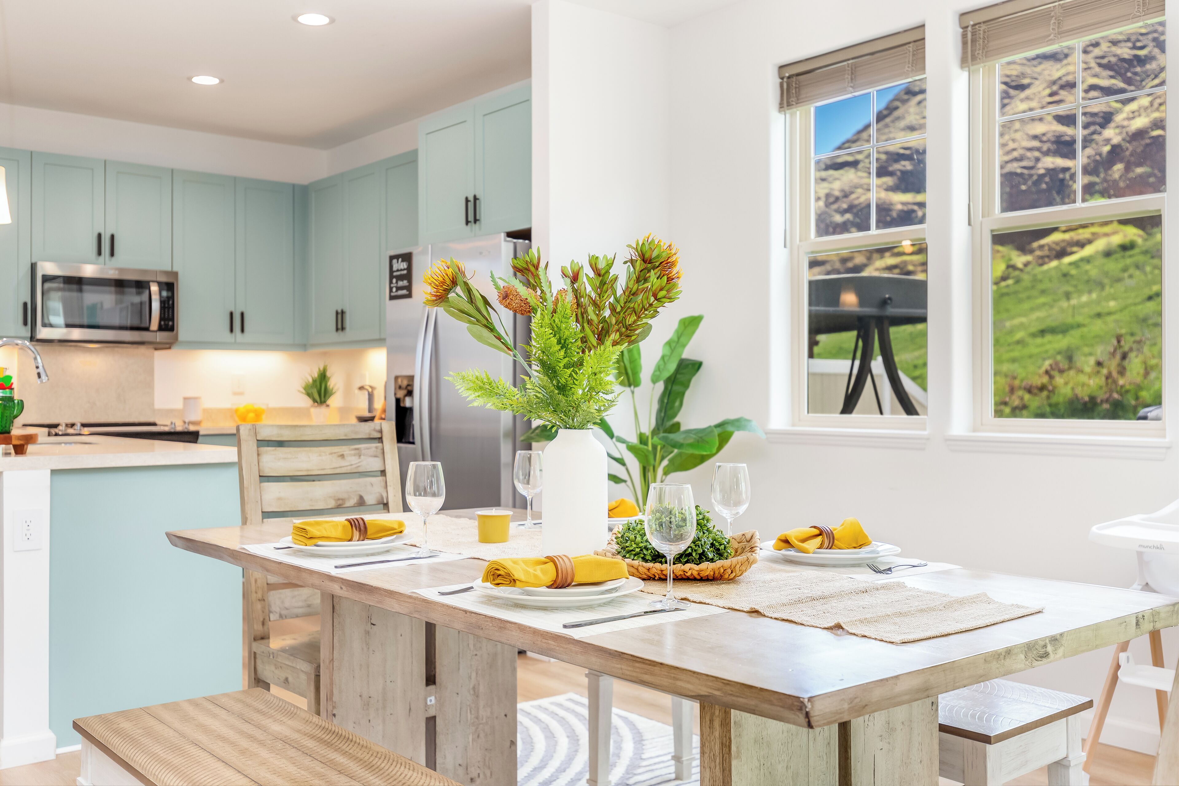 Una cocina moderna con armarios color aguamarina, una mesa de comedor de madera con servilletas amarillas y vistas a un paisaje montañoso a través de amplios ventanales.