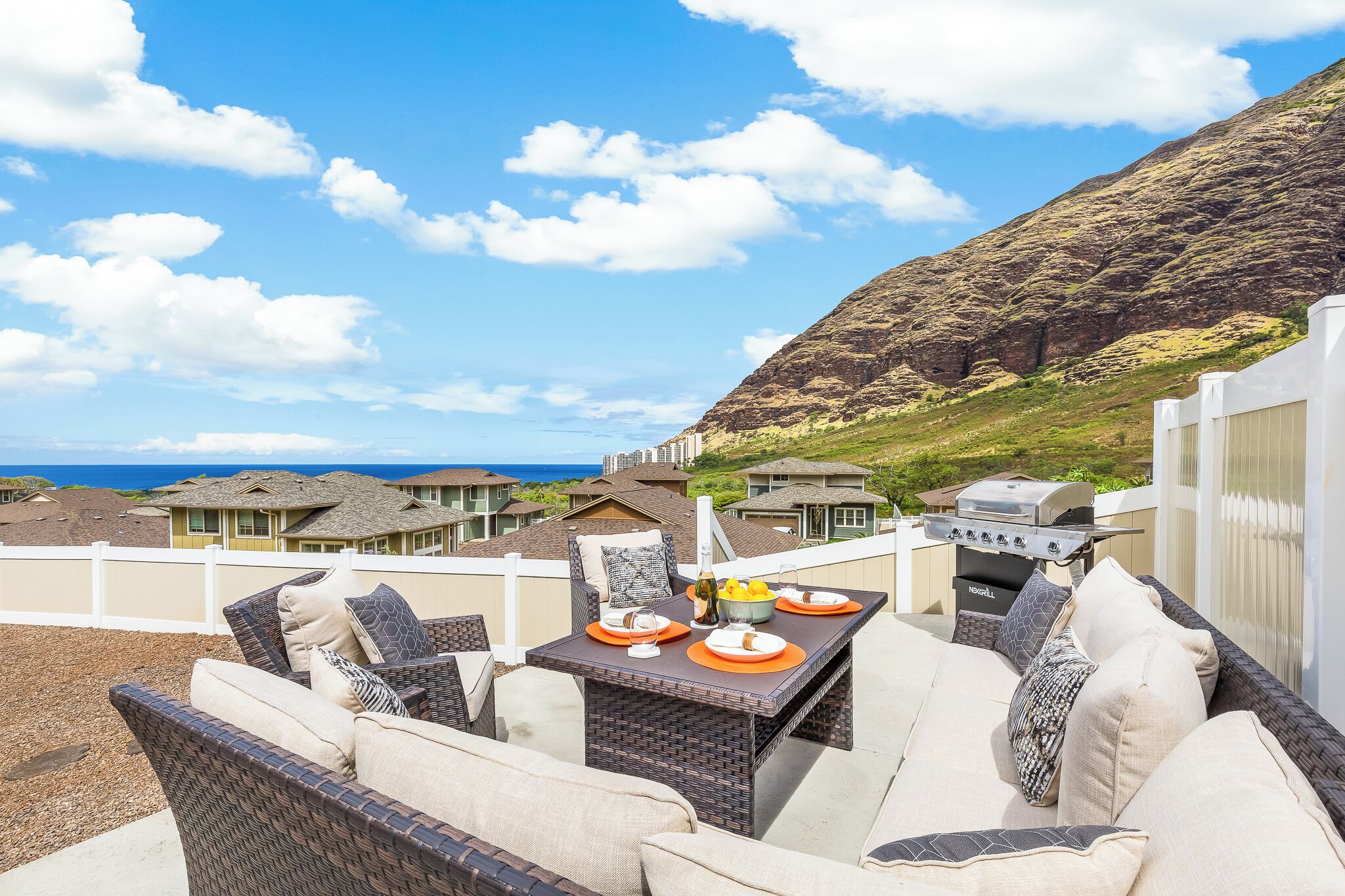 Un área de estar al aire libre con mobiliario de mimbre y una mesa lista para comer, con vistas a un paisaje montañoso y el océano.