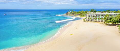 A beach with clear turquoise waters, a sandy shore, and a resort building with multiple floors and balconies overlooking the ocean.