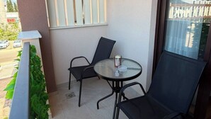 Un balcone con un tavolino in vetro e due sedie, una finestra con persiane e vista su un edificio.