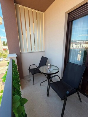 A balcony with a glass table and two chairs, a window with blinds, and a view of a building. - Peaceful 1-bedroom apartment with AC in charming Podgorica (Podgorica)