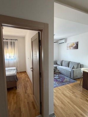 A room with a wooden floor, a grey sofa, a small table, and a chair. - Peaceful 1-bedroom apartment with AC in charming Podgorica (Podgorica)