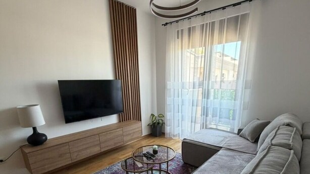 Un soggiorno moderno con divano, tavolino, televisore su un mobile in legno e una grande finestra con tende.