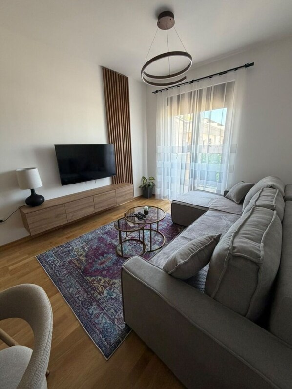 A modern living room with a sofa, a coffee table, a TV on a wooden cabinet, and a large window with curtains. - Peaceful 1-bedroom apartment with AC in charming Podgorica (Podgorica)