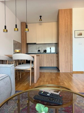 A modern living room with a sofa, a coffee table with remotes, and a kitchen area with wooden cabinets and a countertop. - Peaceful 1-bedroom apartment with AC in charming Podgorica (Podgorica)