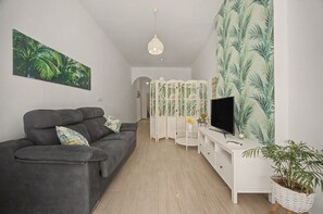 Ein modernes Wohnzimmer mit einem grauen Sofa, einem Fernseher auf einem weißen Schrank, einer Zierpflanze und einer Wand mit tropischem Dekor.