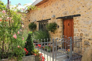 A stone building with a wooden door, a metal gate, and a garden with roses and shrubs.