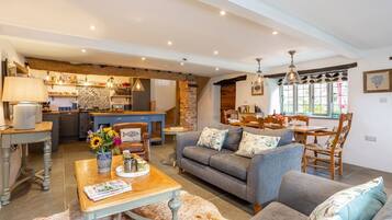 A spacious living area with a sofa, coffee table, and dining table. There is a kitchen in the background with a brick wall and pendant lights.