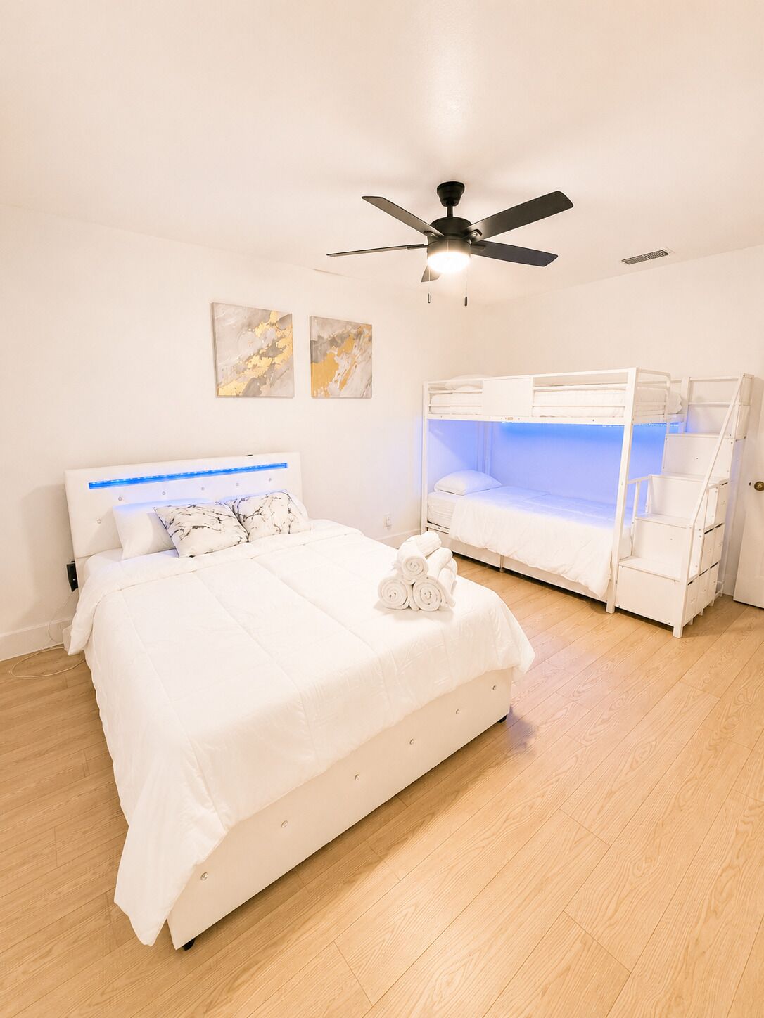 A bedroom with a large bed, a bunk bed, a ceiling fan, and artwork on the wall.