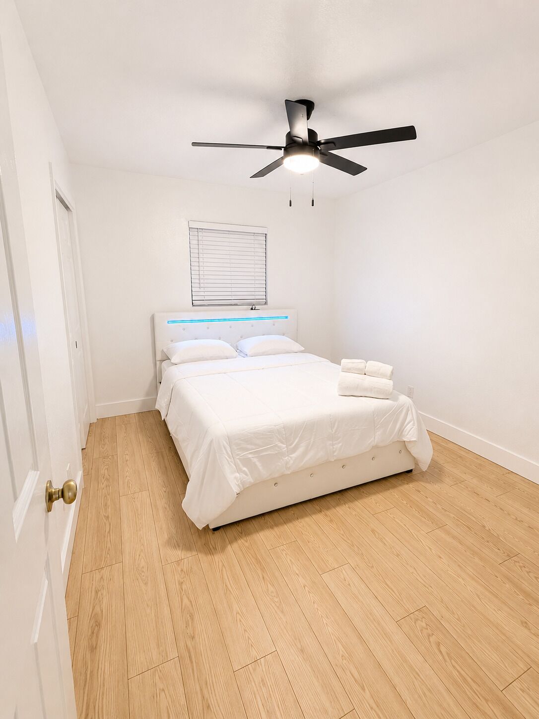 A bedroom with a bed, a ceiling fan, a window with blinds, and a wooden floor.