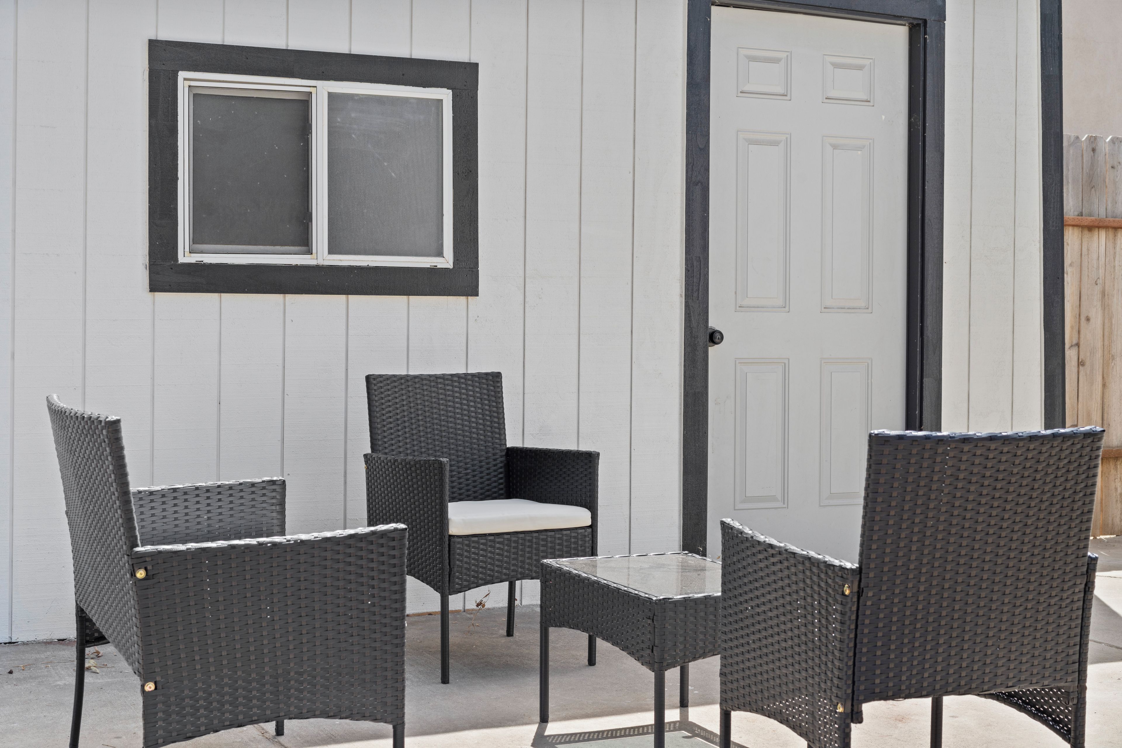 Een terras met twee rieten stoelen en een kleine tafel, een witte deur en een raam met hor.