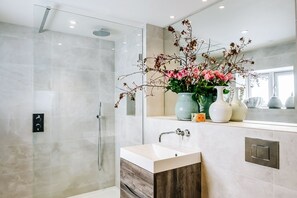 Un bagno moderno con doccia vetro, lavabo con mobiletto in legno e vasetto di fiori sul piano di lavoro.