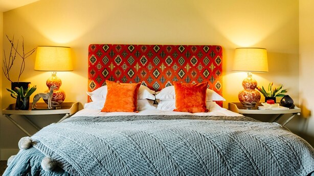 A hotel room with a patterned headboard, two bedside lamps, a bed with white linens and orange pillows, and a blue knitted throw.