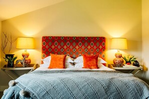Una camera d'albergo con una testiera a motivi decorativi, due lampade ai lati del letto, un letto con lenzuola bianche e cuscini arancioni, e una coperta lavorata a maglia blu.
