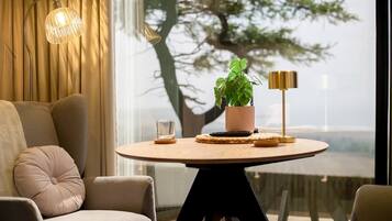 A modern living room with a round table, a sofa, a potted plant, a lamp, and a view of a tree through a window.