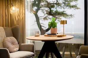 Ein modernes Wohnzimmer mit einem runden Tisch, einer Couch, einer Topfpflanze, einer Lampe und Blick auf einen Baum durch ein Fenster.