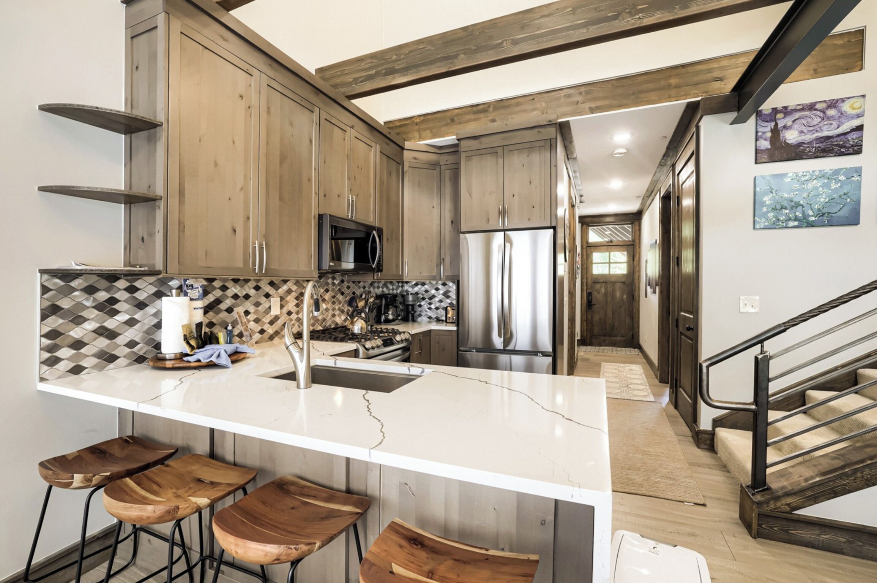 Una cocina moderna con una isla central, taburetes de bar, armarios de madera y electrodomésticos de acero inoxidable, además de una escalera que conduce a otro nivel.