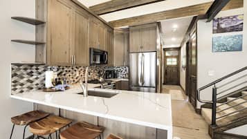 Una cocina moderna con una isla central, taburetes de bar, armarios de madera y electrodomésticos de acero inoxidable, además de una escalera que conduce a otro nivel.