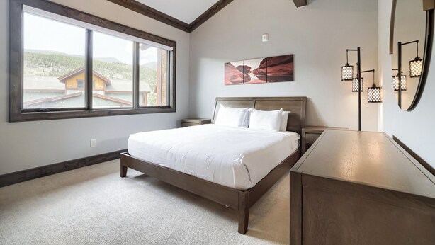 Une chambre avec une grande fenêtre, un lit, une table de chevet, une commode, un tableau et un miroir.