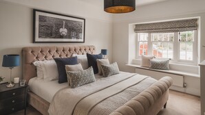Quarto com cabeceira estofada, cama grande com várias almofadas, mesa de cabeceira com abajur e janela com vista para edifícios.