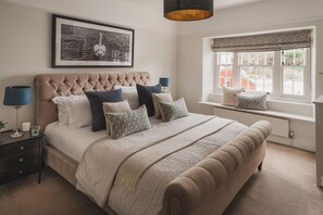 Een slaapkamer met een gebogen hoofdeinde, een groot bed met veel kussens, een nachtkastje met een lamp en een raam met uitzicht op gebouwen.