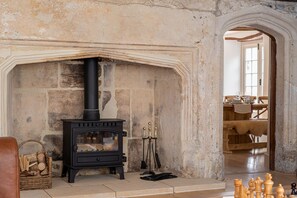 A stone fireplace with a black wood stove, a basket of firewood, and a chessboard on the floor.